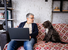 Man sat on red couch with laptop on lap next to a brown dog